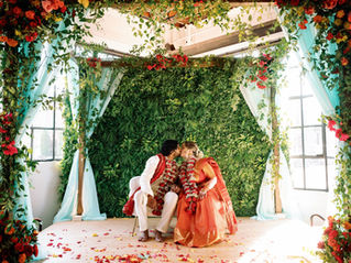 Bride and groom sitting under a mandap, or altar, at a cultural wedding. The altar consists of four pillars covered in drapery and colorful florals with greenery.