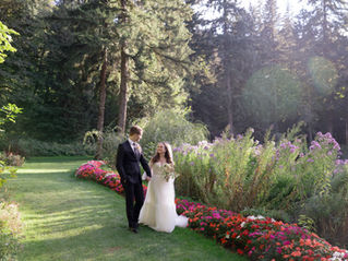 Bride and groom walk hand in hand down a grass path lined with colorful florals and towering trees behind them.