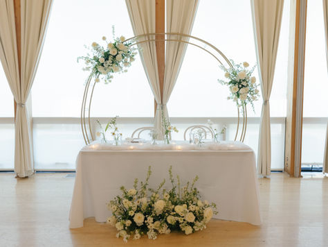 Sweetheart table topped with bud vases with a variety of white florals, a large white floral arrangement at the foot of the table, and a large circular gold arch with two more white floral arrangements attached behind the table.