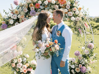 Bride and groom sharing a loving look at the altar in front of a large, beautiful floral arrangement with vineyard views to complete the backdrop.