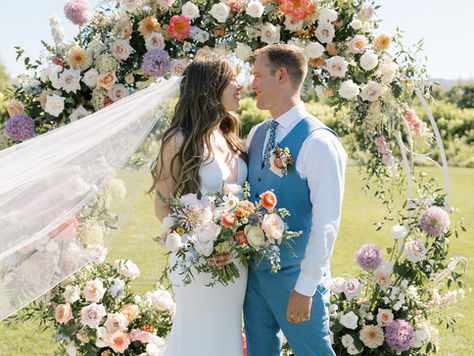Bride and groom sharing a loving look at the altar in front of a large, beautiful floral arrangement with vineyard views to complete the backdrop.