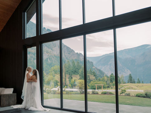 Two beautiful brides embracing each other in front of a wall of windows that look out on a gorgeous mountain range view in the Columbia River Gorge.