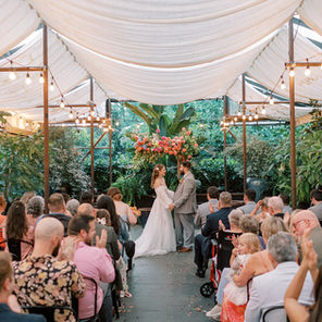 Bride and groom standing at the altar with a bright floral arrangement behind them on the arbor, surrounded by greenery, and under the cover of cafe lights and drapery. Friends and family watch as they say their vows.