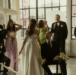 This photo shows a wedding party gathered inside Castaway Portland, a bright industrial venue with tall black-framed windows that let in soft natural light. The group is arranged casually throughout the space, some holding white parasols and others holding elegant white flowers, creating an artistic scene