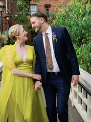 Bride in a fun chartreuse dress walking arm in arm with her groom, sharing a loving glance, down a fenced path surrounded by greenery.