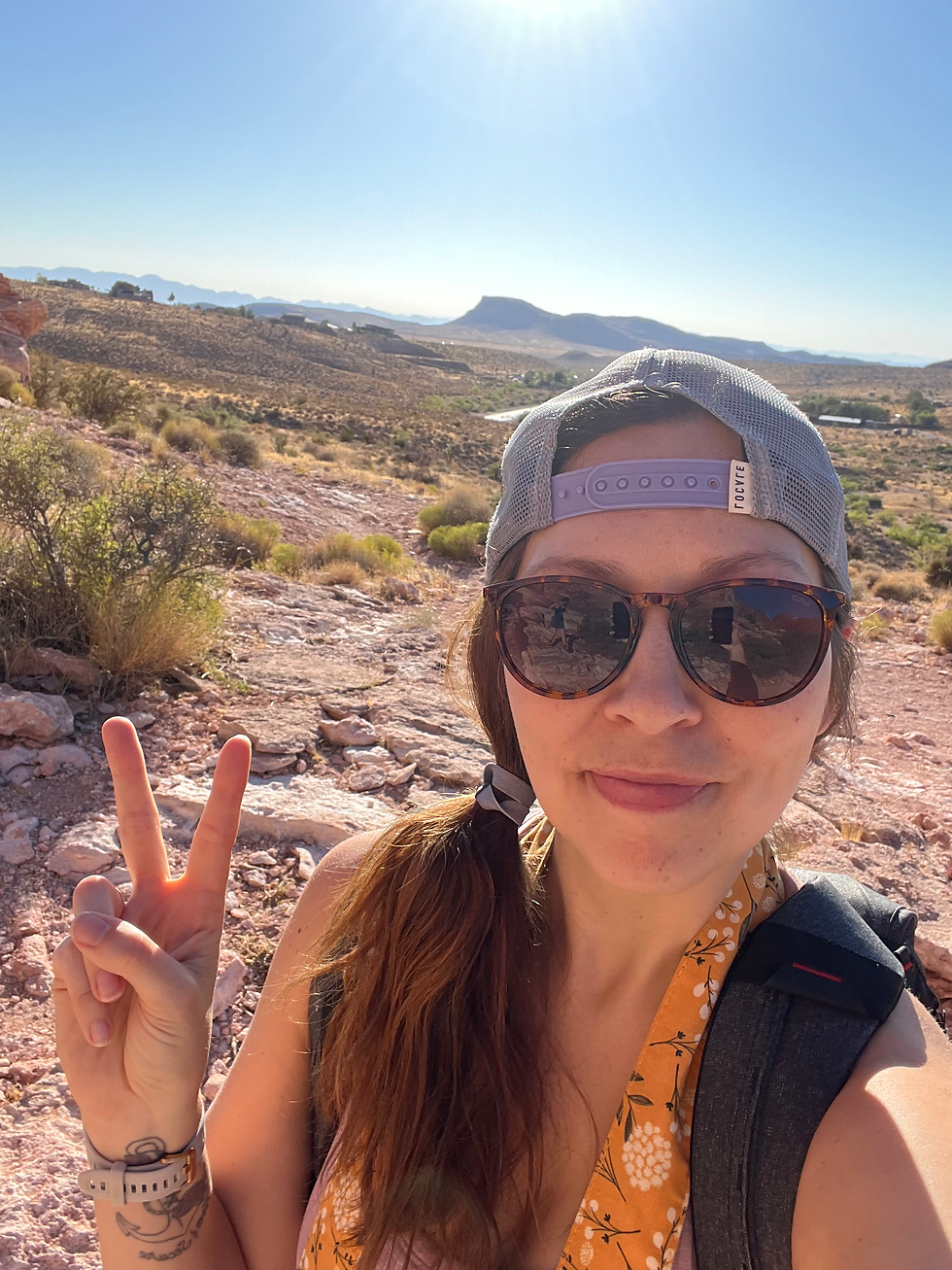 Woman in sunglasses and cap gives peace sign in desert landscape. Bright sun overhead, wearing floral top, looks content.