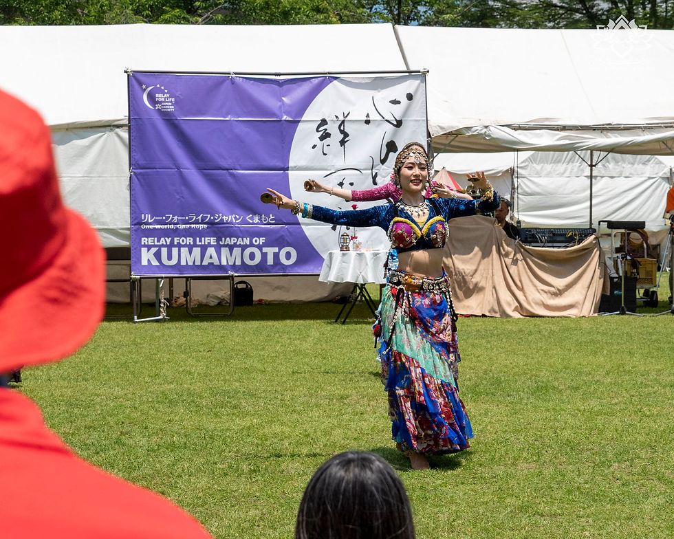 芝生の屋外ステージで、民族衣装の女性ダンサーが腕を広げて踊る。背後に「RELAY FOR LIFE JAPAN OF KUMAMOTO」の紫の幕。