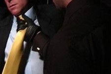 A man in a suit and tie, bound and captive, submits to a dark figure with a gloved hand.