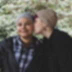Two people in hand knit hats outdoors; one kisses the other's cheek as they smile softly against leafy greenery. The pattern was designed by Wool & Wyrd.
