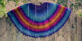 Colorful knit shawl draped over wooden fence, featuring vibrant rainbow stripes. Lush greenery and rustic wooden planks add natural texture.