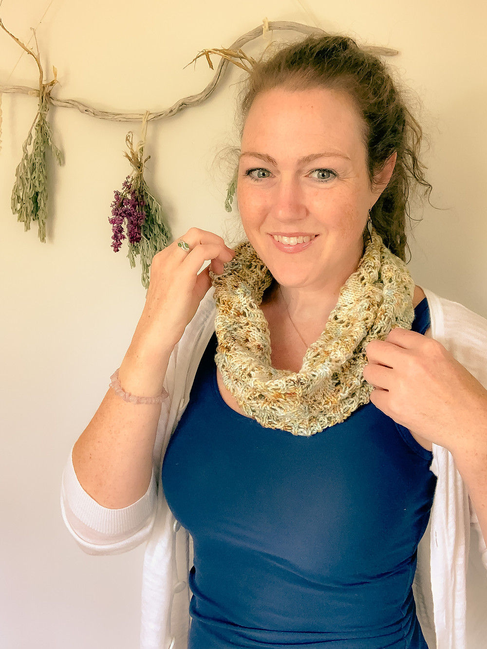 A white lady stands infront of drying herbs. She is wearing a hand knit lace cowl.