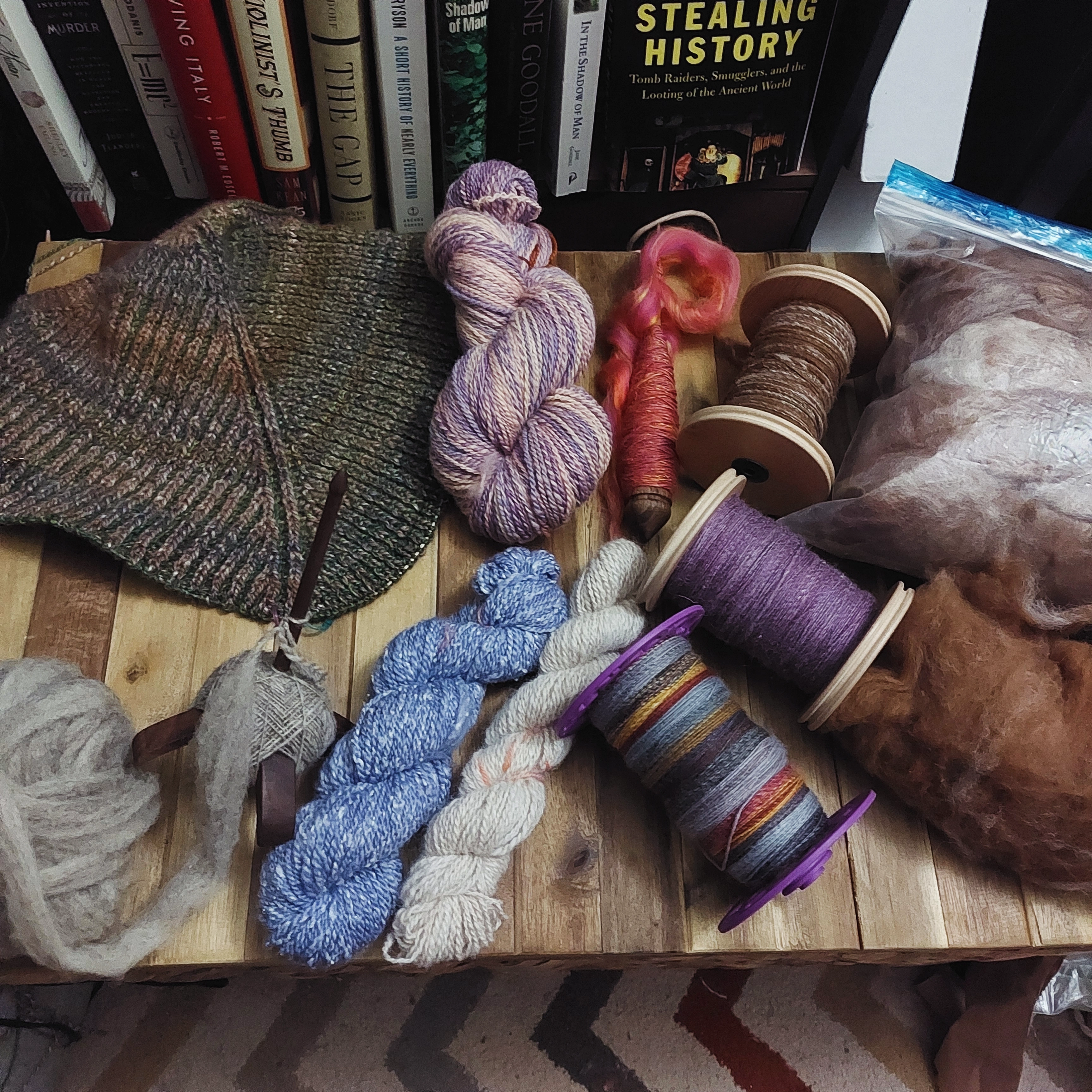 Various colorful hand spun yarns, bobins, spindles, and roving are spread on a wooden table. Books and a rug are visible in the background. Cozy and creative vibe.