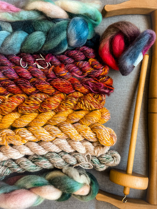 Colorful hand spun yarn skeins and roving wool lie on a gray surface beside a wooden spindle, showcasing vibrant reds, yellows, and greens.