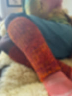 Person with a cozy style, multicolored sweater relaxes on a fluffy surface. They're wearing hand knit orange leg warmers and pink socks. Bird art on wall.