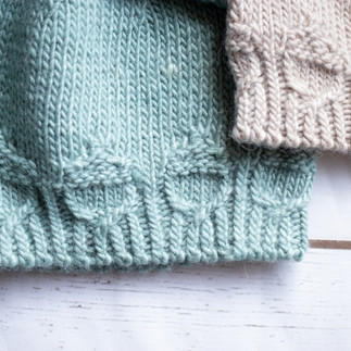 Close-up shot of a grey knit beanie's brim showing intricate cable stitches that look like tiny acorns, resting on a white wood background. Pattern by Wool & Wyrd.