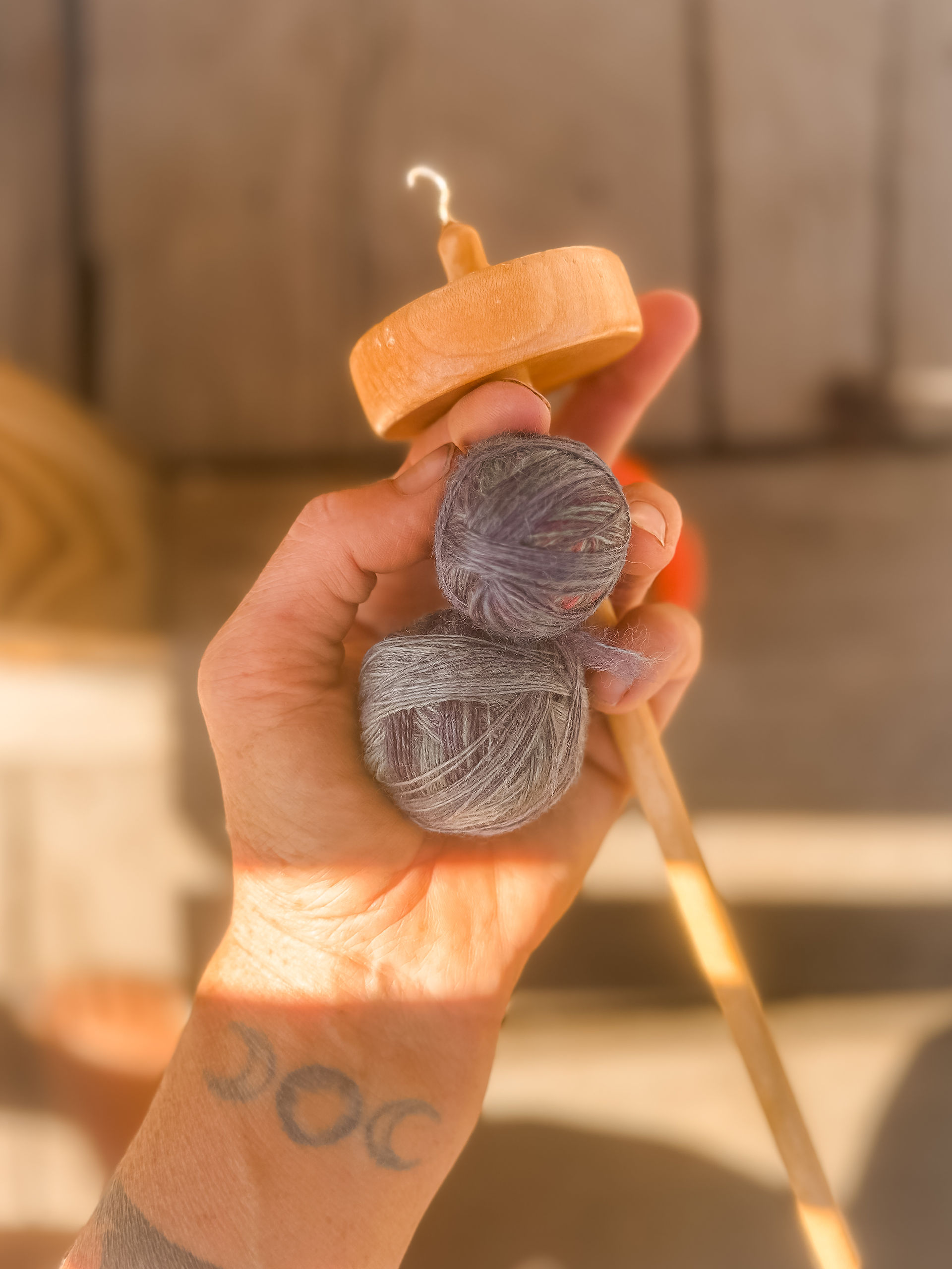 Hand holding a drop spindle and gray yarn balls, with a crescent moon tattoo on wrist. Sunlit wooden background creates a warm mood.