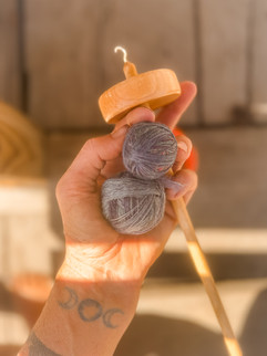 Hand holding a drop spindle and gray yarn balls, with a crescent moon tattoo on wrist. Sunlit wooden background creates a warm mood.