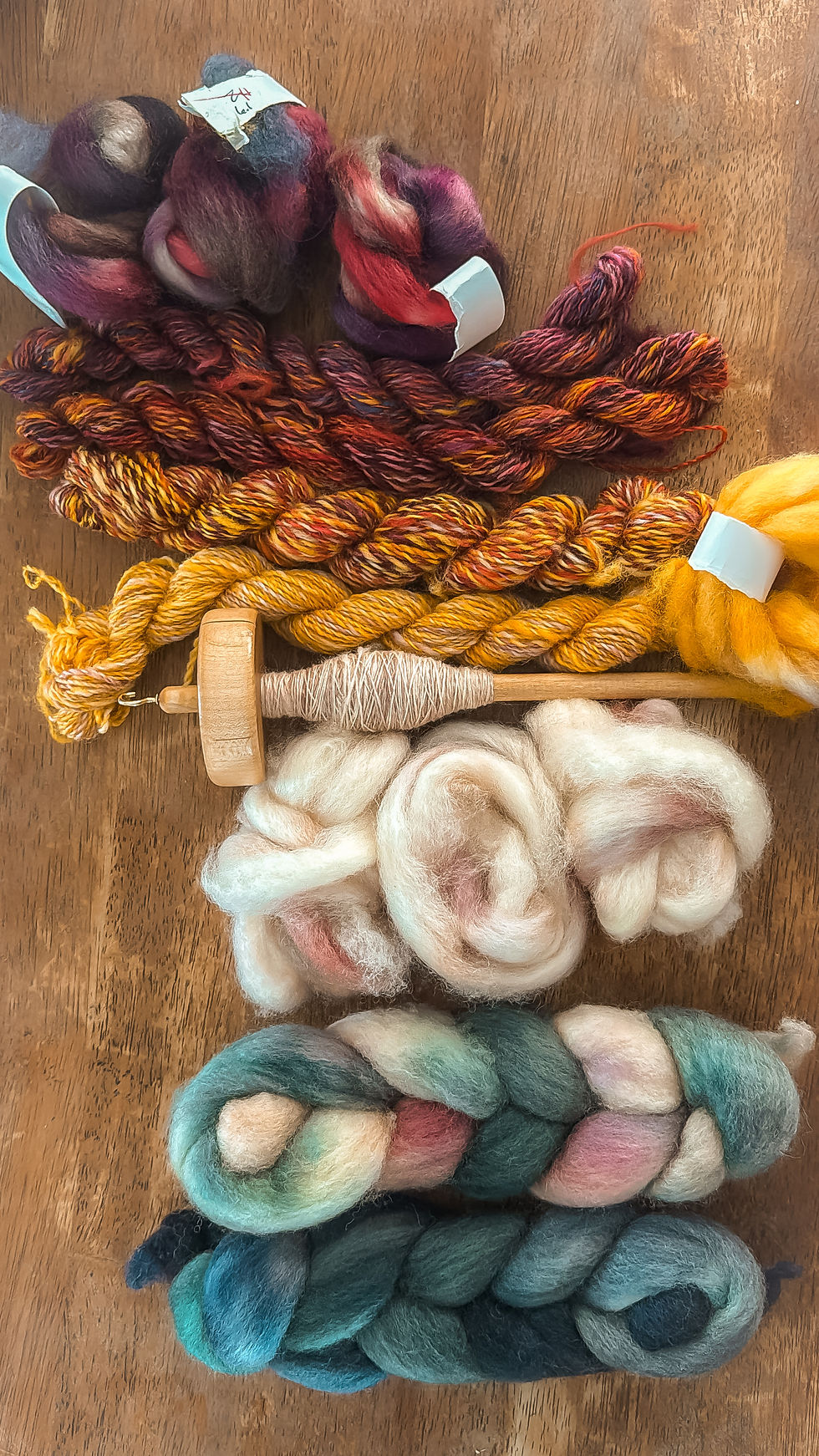 Colorful yarn skeins and fibers, including a spindle and wool, on a wooden table. Varied hues of red, yellow, and teal are visible.
