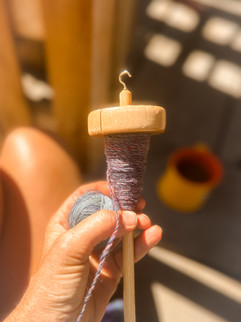 Hand spins purple hand spun yarn on a wooden spindle in sunlight. A ball of yarn is held in the other hand. Sunlit wooden background.