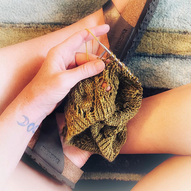 A hand holds a lace cowl on the knitting needles, mid stitch. The yarn and cowl are green, the hand and lap are a white lady's and the feeling is soft and comfortable.