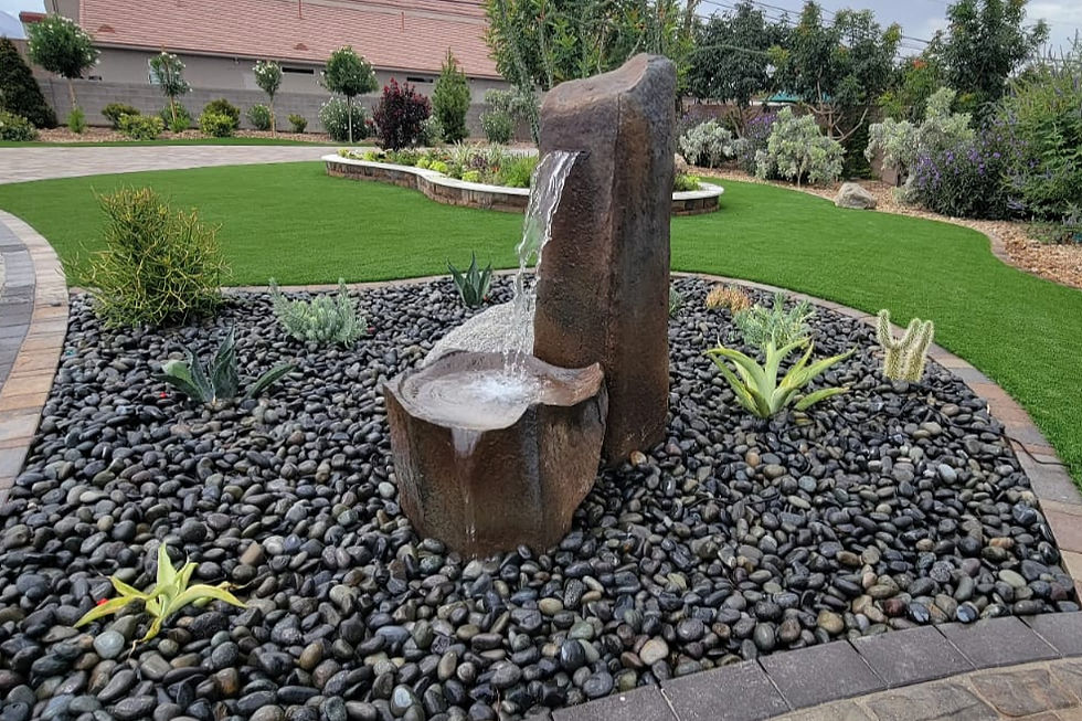 A crisp garden scene featuring a decorative stone fountain, with water cascading gently onto a bed of smooth black pebbles, surrounded by vibrant green plants and manicured grass.