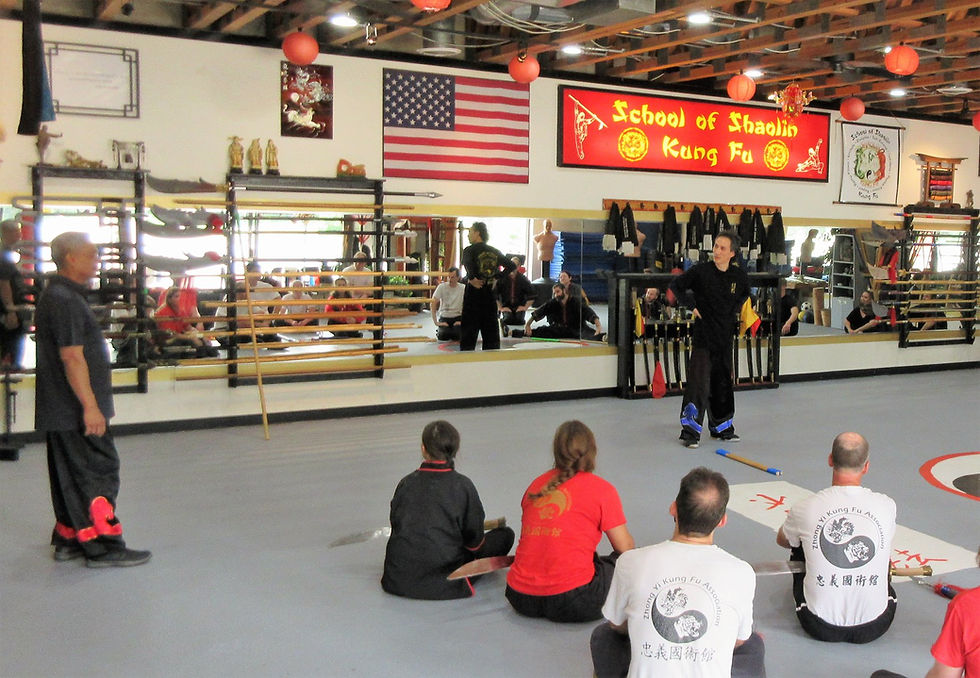 A martial arts instructor Rick "Bucky" Wing teaches seated students at the School of Shaolin Kung Fu. Mirrors, weapons, and an American flag adorn the dojo.