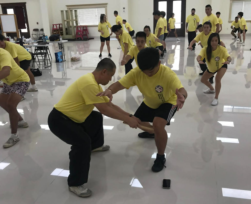 People in yellow shirts practice lion dance in a bright room. Two individuals are focused in the foreground, crouching with a stick.
