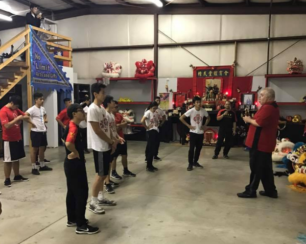 Group of people practicing lion dance in a decorated studio. A man in red instructs. Lion dance props and a blue banner in the background.