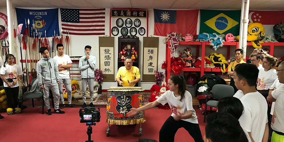Drumming demonstration with Grandmaster Chan Siew Kee in a decorated room with flags and martial arts gear. Participants watch and film. Energetic mood.