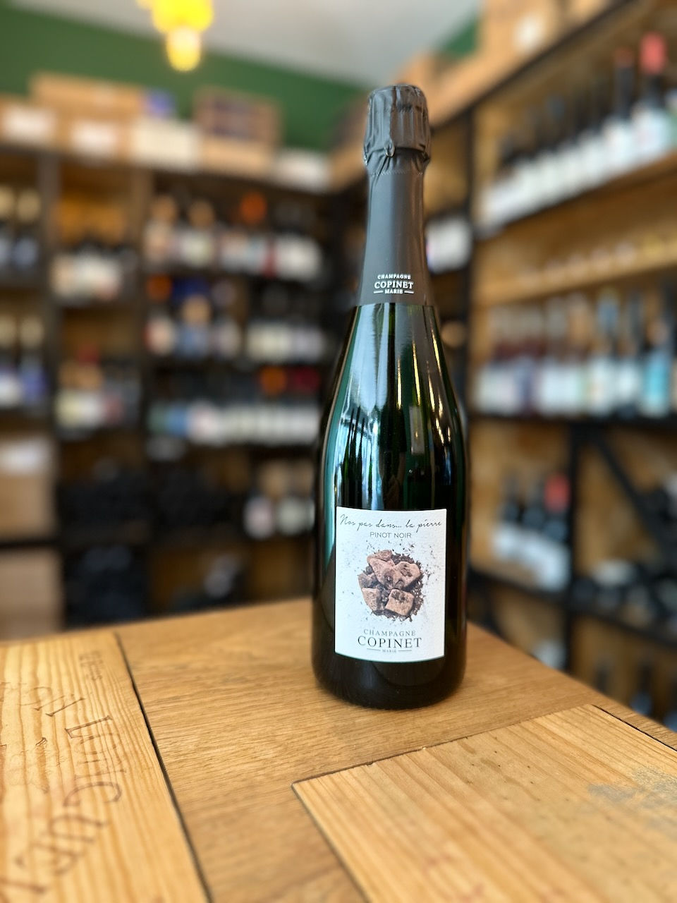 bouteille de champagne Nos pas dans la craie 100% Pinot noir du Domaine Marie Copinet posée sur une table en bois devant des étagères remplies de bouteilles de vins
