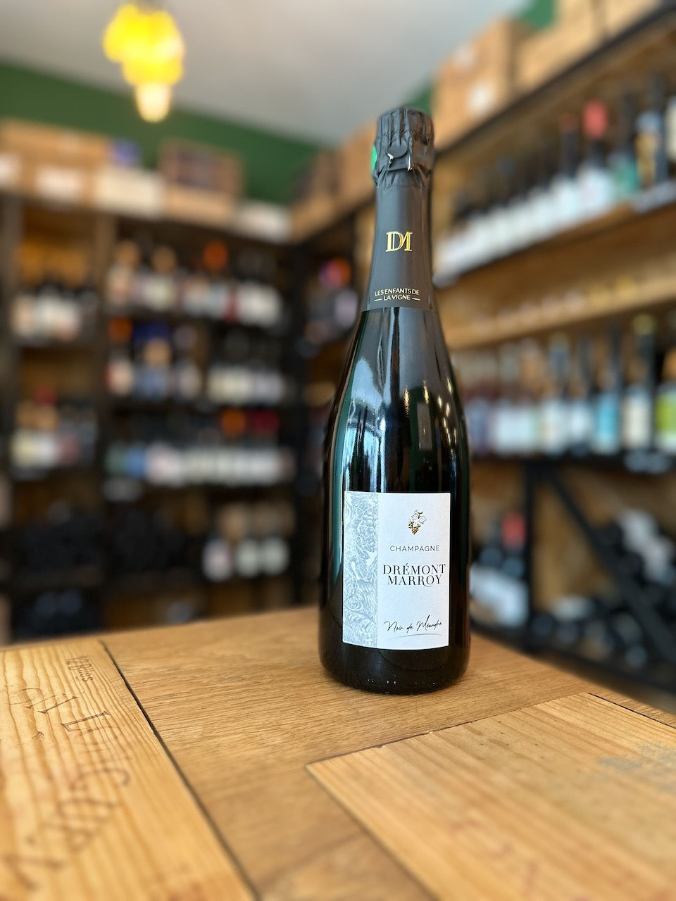 cuvée champagne Noir de Méandres du domaine Drémont Marroy posée sur une table en bois devant des étagères remplies de bouteilles de vins