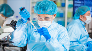 A male scientist in a blue lab gown and mask testing a glass of water.