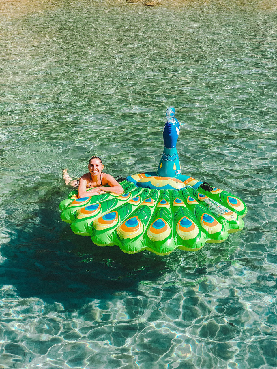 solo female traveler floating on a peacock pool float in shallow turquoise waters