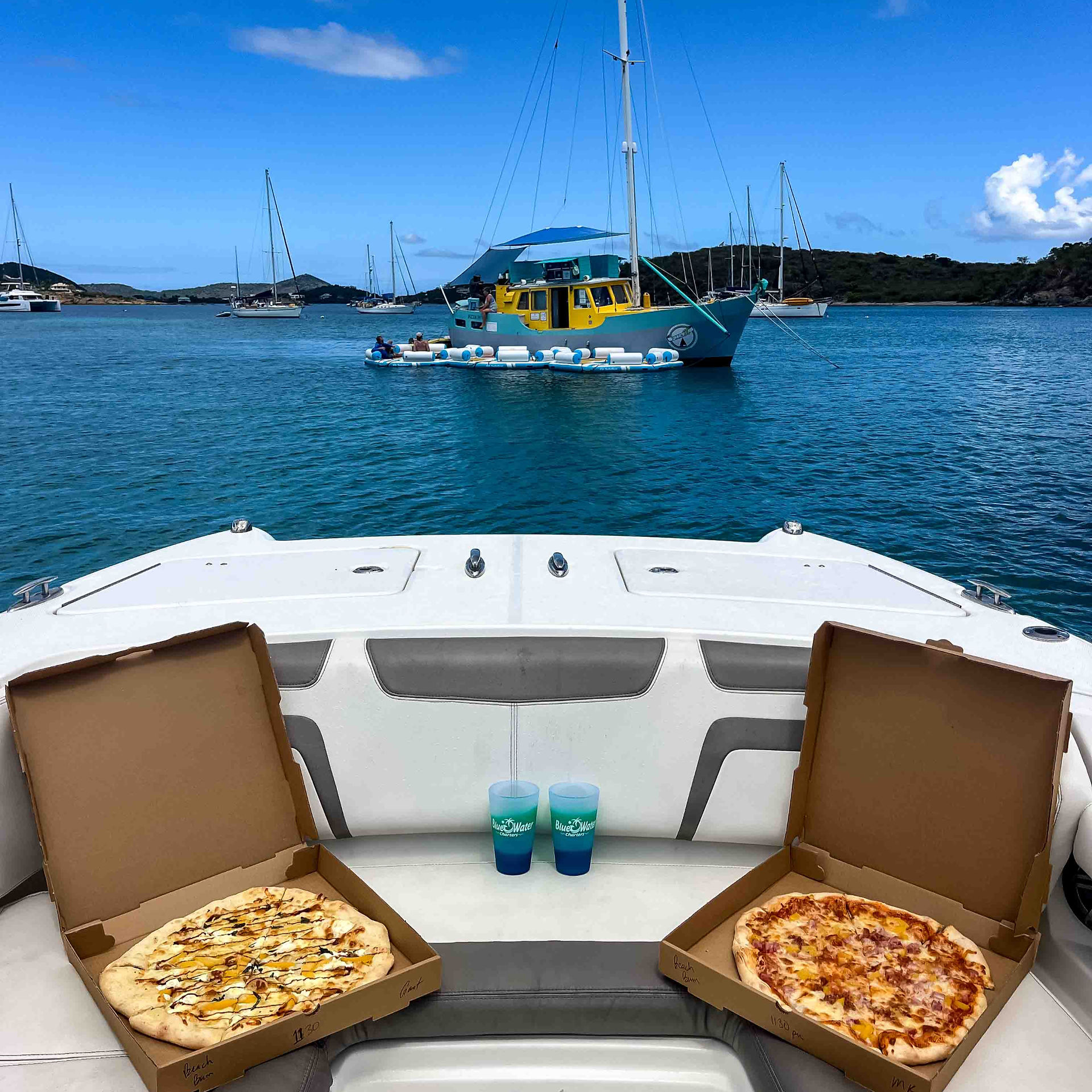 Two pizzas and tropical drinks on the bow of a boat with Pizza Pi floating pizzeria in the background – take-out, Virgin Islands style.