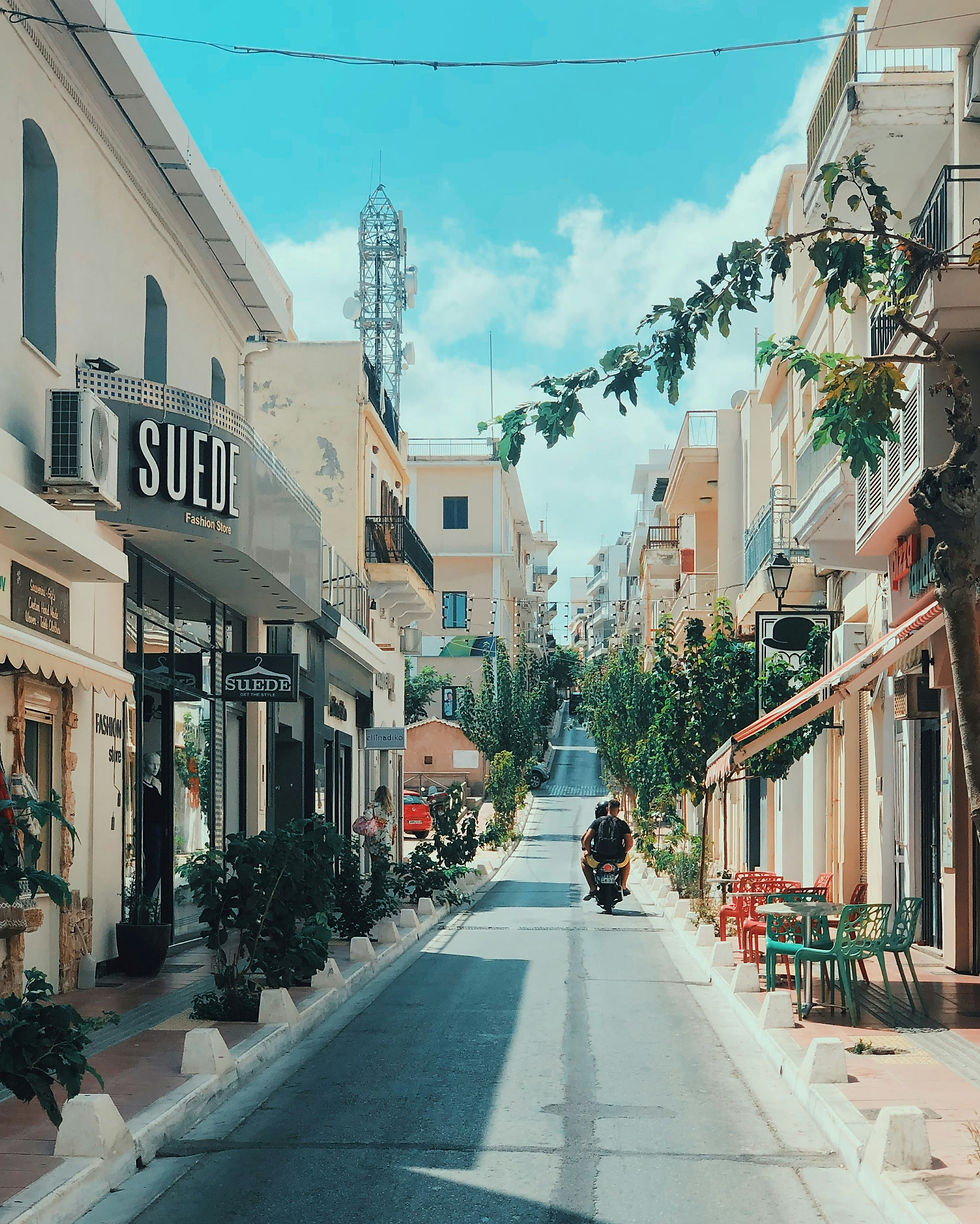 Sunny narrow street lined with buildings and plants. A person rides a scooter toward the horizon. "SUEDE" sign on a shop. Calm mood.