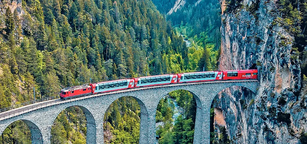Red train crosses stone viaduct in lush forested mountains. Bright and serene setting with vibrant greenery. "Glacier Express" text visible.