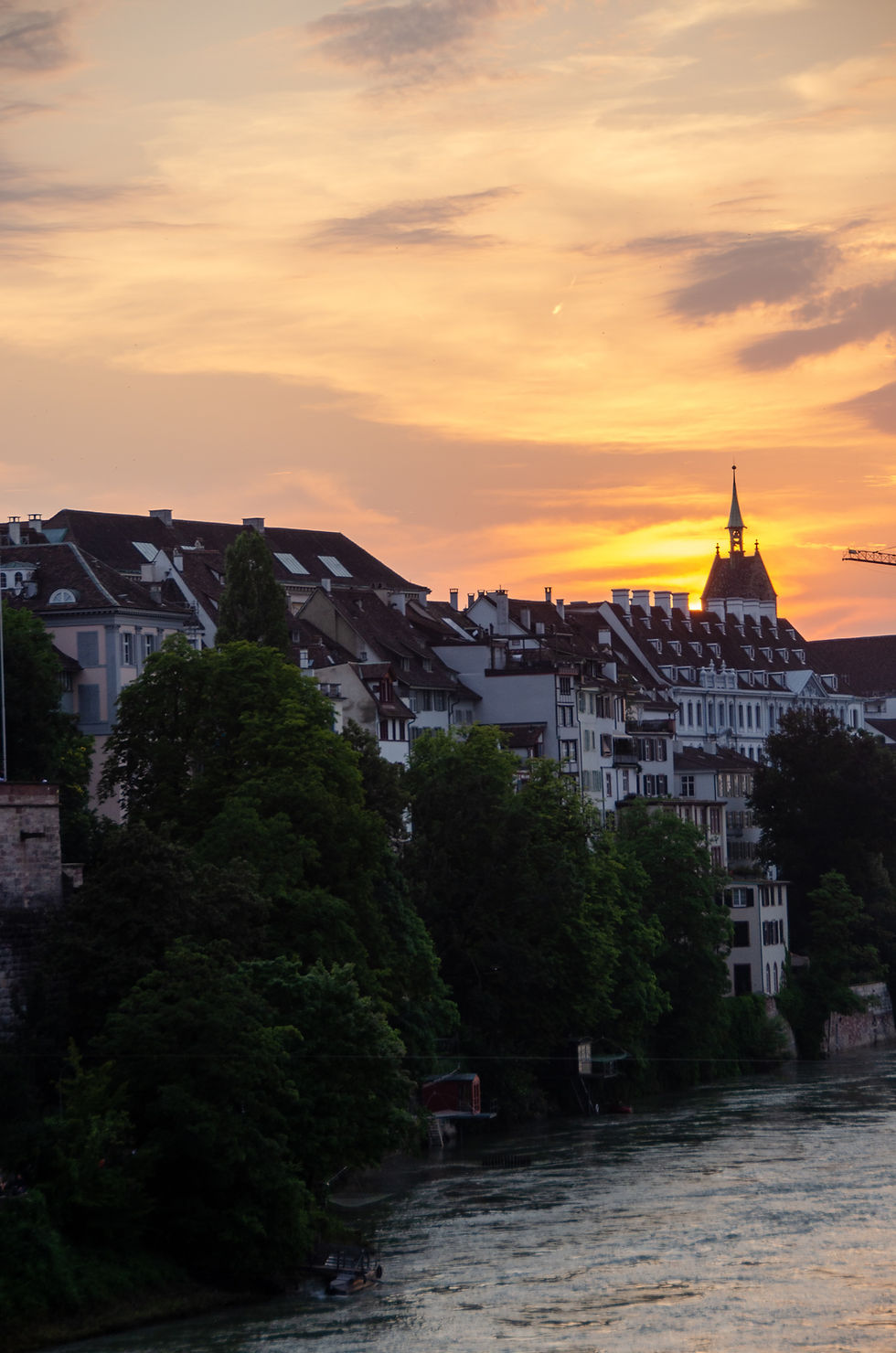 Riverside European town at sunset, featuring picturesque houses with steep roofs, lush trees, and a vibrant orange sky. Peaceful ambiance.