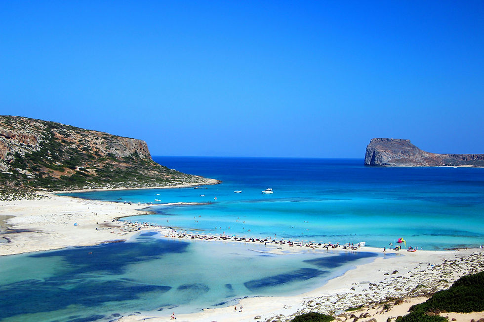 Beach scene with people relaxing on white sand and swimming in turquoise water. Boats float nearby, with rocky cliffs and clear blue sky.