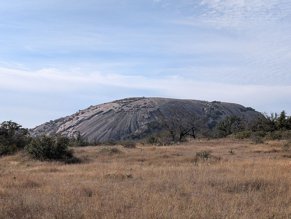 Rocky hill under a clear blue sky with scattered clouds, surrounded by dry, grassy terrain and sparse trees. Peaceful, natural setting.