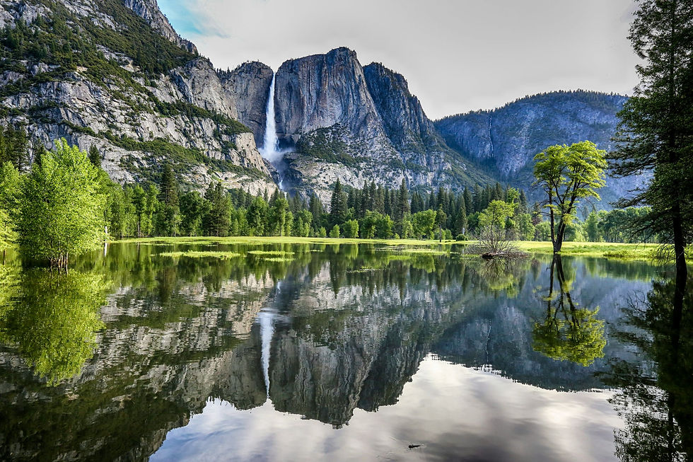 Waterfall cascades down a rocky mountain into a lush green valley with trees reflecting in a calm lake under a partly cloudy sky.