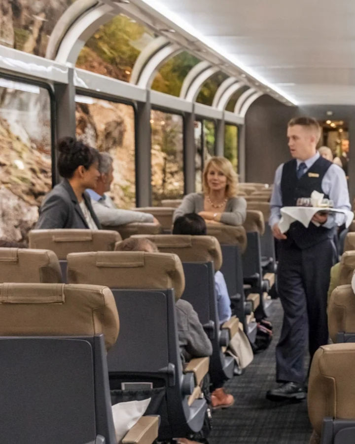 Passengers sit and chat in a train car with large windows. A server in a vest carries a tray. The mood is relaxed and sociable.