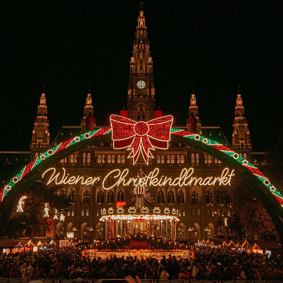 Crowds gather at Vienna's Christmas market under colorful lights and arch with "Wiener Christkindlmarkt" sign. Festive and vibrant scene.