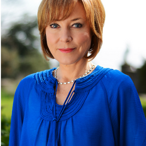 Sian Williams in blue blouse with short brown hair stands outdoors, looking at the camera. Blurred green background, clear sky. Calm expression.