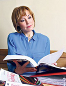 Sian Williams in a blue shirt reads a book, surrounded by colourful magazines. She appears focused in a simple indoor setting with a beige wall.