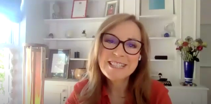 Sian Williams smiling woman in glasses with earbuds, sits in a bright room with white shelves, framed art, and a vase of flowers. She wears a red shirt.