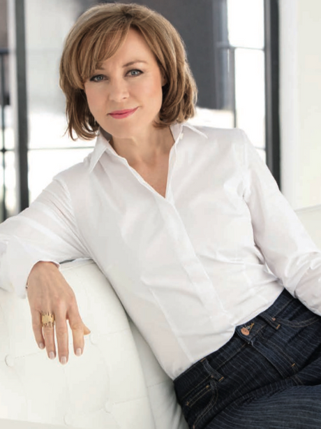 Sian Williams in white blouse and jeans leaning on a white sofa in a bright room. She appears relaxed and confident, with a soft smile.