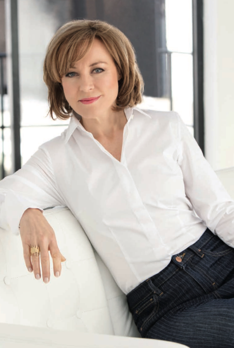 Sian Williams in white blouse and jeans leaning on a white sofa in a bright room. She appears relaxed and confident, with a soft smile.
