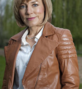 Sian Williams in a brown leather jacket smiles outdoors against a green, blurred background. She wears a white shirt and pearl necklace.
