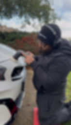 Man polishing white car fender with power tool, wearing beanie, headphones.