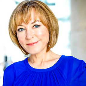 Sian Williams in bright blue top smiling, standing between stone columns. Background is blurred, with natural light illuminating her face.